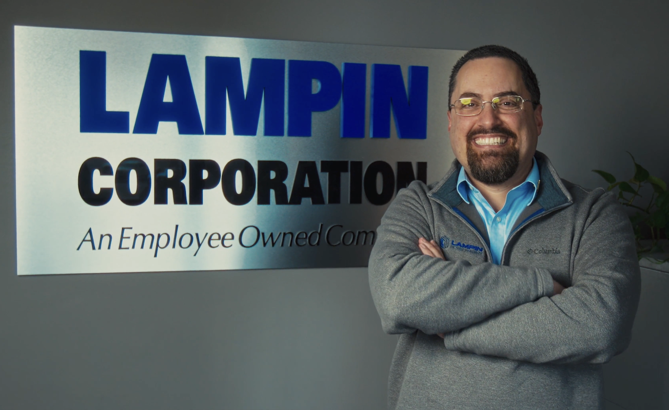 John Biagioni, a man with light skin, dark brown hair, glasses, and a brown beard, stands in front of a Lampin sign. He is smiling proudly and stands in a confident pose with his arms crossed in front of him.