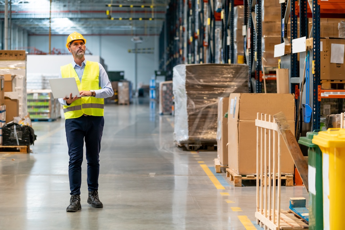 A tall man in a yellow vest and hardhat carries a laptop computer through the warehouse area of a factory.